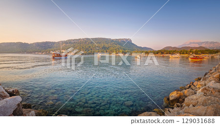 Morning light bathes the Moonlight beach in Kemer, Turkey. Boats rest in the calm bay with turquoise Mediterranean sea water. Lush green hills with palm trees seen at horizon Morning light bathes the Moonlight beach in Kemer, Turkey. Boats rest in the calm bay with turquoise Mediterranean sea water. Lush green hills with palm trees seen at horizon 129031688