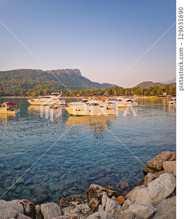 Morning light bathes the Moonlight beach in Kemer, Turkey. Boats rest in the calm bay with turquoise Mediterranean sea water. Lush green hills with palm trees seen at horizon 129031690