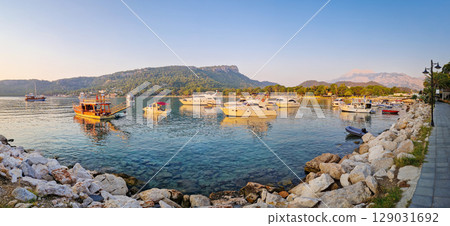 Morning light bathes the Moonlight beach in Kemer, Turkey. Boats rest in the calm bay with turquoise Mediterranean sea water. Majestic Taurus mountains with Olympos peak Tahtali in the background Morning light bathes the Moonlight beach in Kemer, Turkey. Boats rest in the calm bay with turquoise Mediterranean sea water. Majestic Taurus mountains with Olympos peak Tahtali in the background 129031692