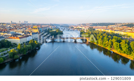 Aerial view shows the Vltava River winding through Prague, with the National Theatre on the banks. Lush green trees line the waterfront under a clear sky, highlighting the city's beauty. 129031809
