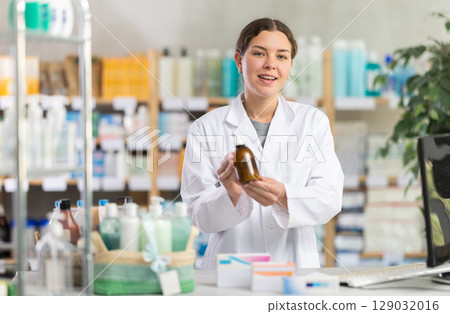 Professional young girl pharmacist standing at prescription counter and offering medicinal cough liquid syrup to clients at pharmacy store Professional young girl pharmacist standing at prescription counter and offering medicinal cough liquid syrup to clients at pharmacy store 129032016