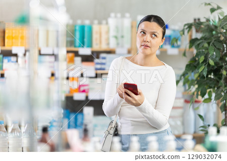 Middle-aged woman scrolling phone while looking for something in drugstore Middle-aged woman scrolling phone while looking for something in drugstore 129032074