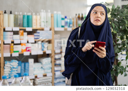 Middle-aged Muslim woman scrolling phone while looking for something in drugstore Middle-aged Muslim woman scrolling phone while looking for something in drugstore 129032318