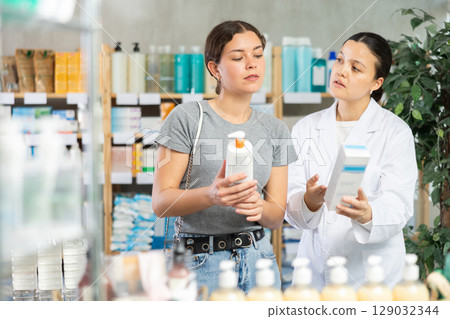 Focused young girl choosing cream or skin care product in pharmacy, young asian woman pharmacist consulting and offering variety of goods to customer 129032344