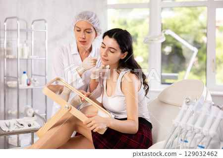 Cosmetologist showing results of facial procedure to young woman Cosmetologist showing results of facial procedure to young woman 129032462