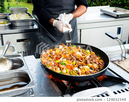 Male chef cooking stir fry outdoors with colorful vegetables and meat in a pan Male chef cooking stir fry outdoors with colorful vegetables and meat in a pan 129032549