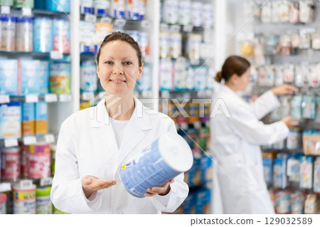 Portrait of female pharmacist in sales hall of a pharmacy, demonstrating jar of baby food 129032589