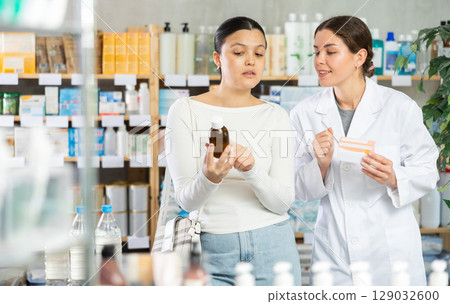 Asian woman buyer choosing medicinal cough liquid syrup standing between shelves at pharmacy, young girl pharmacist consulting customer Asian woman buyer choosing medicinal cough liquid syrup standing between shelves at pharmacy, young girl pharmacist consulting customer 129032600