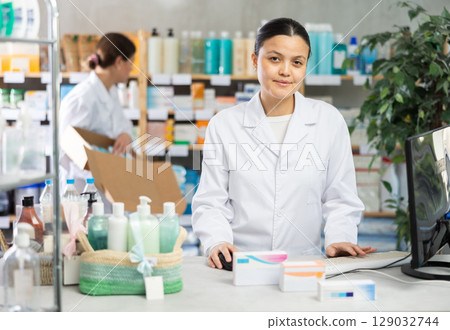 Middle-aged female pharmacist working with computer and other restocking in background Middle-aged female pharmacist working with computer and other restocking in background 129032744
