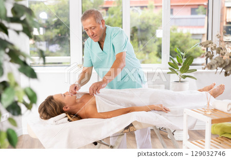 Kind aged masseur covering woman lying on massage table with blanket 129032746