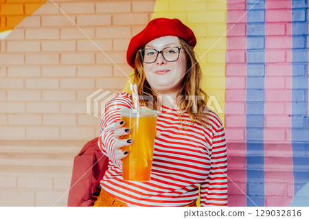 Smiling plus size woman in glasses and bright clothes holding plastic glass with sugar flavored tapioca bubble tea near urban colorful painted brick wall. 129032816