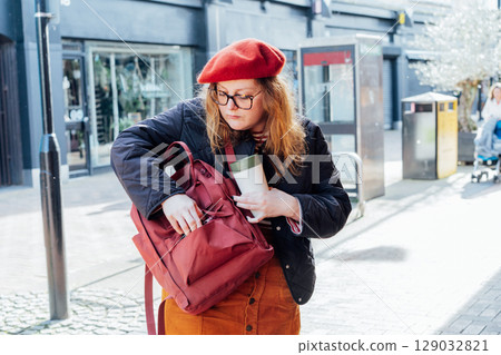 A young woman in a red beret and casual attire searching something in her burgundy backpack and holding reusable coffee cup while walking down a lively city street lined with shops on a sunny day 129032821