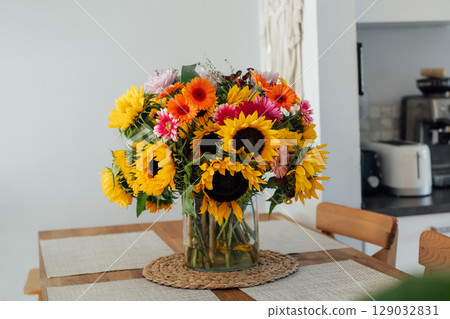 Stylish modern interior of open space white kitchen with huge multicolor summer flower bouquet in vase on wooden kitchen counter table, macrame on the wall. Cozy Design home decor. Selective focus. 129032831
