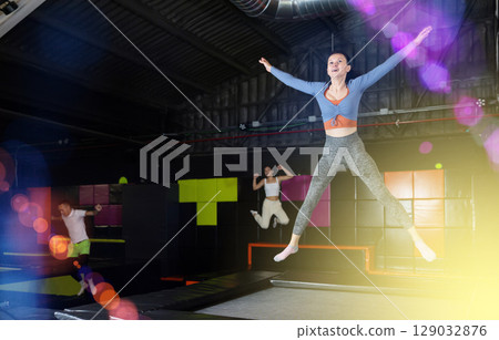 Positive middle aged woman in sporty clothes jumping vertical high in modern trampoline hall 129032876