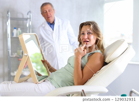 Unsatisfied middle-aged woman holding mirror lying on clinical chair Unsatisfied middle-aged woman holding mirror lying on clinical chair 129032931