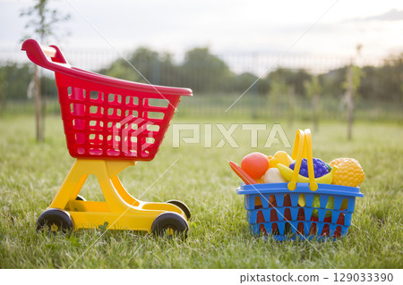 Shopping pushcart and a basket with toy fruits and vegetables. Bright plastic colorful toys for children outdoors on sunny summer day. 129033390