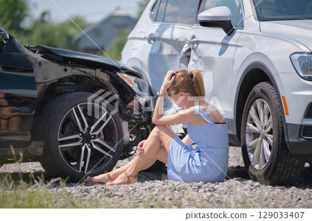 Sad young woman driver sitting near her smashed car looking shocked on crashed vehicles in road accident Sad young woman driver sitting near her smashed car looking shocked on crashed vehicles in road accident 129033407