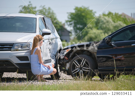Sad female driver sitting on street side shocked after car accident. Road safety and vehicle insurance concept 129033411