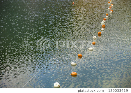 Rope with white and yellow restriction markers on water surface for prohibiting of swimming in deep lake or sea. Rope with white and yellow restriction markers on water surface for prohibiting of swimming in deep lake or sea. 129033419