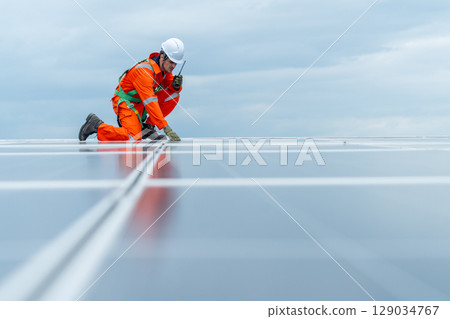 Technician worker or professional engineer sit on solar cell panel and use walkie-talkie during check and maintenance on rooftop of factory building with cloudy sky. Technician worker or professional engineer sit on solar cell panel and use walkie-talkie during check and maintenance on rooftop of factory building with cloudy sky. 129034767