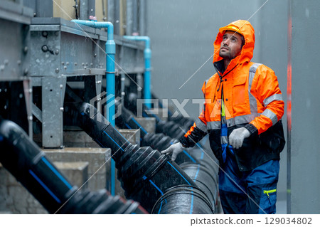 Factory worker or technician with raincoat walk along pipe system and look up to maintenance in area of factory building during raining. 129034802