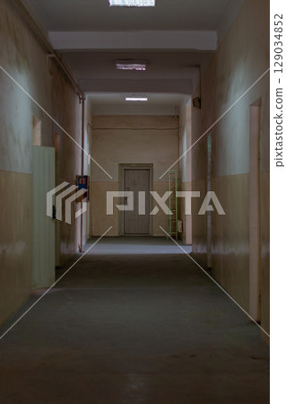 Vertical photo of the empty longitudinal corridor of an old industrial building with high ceilings, illuminated along the entire length. Technical rooms, door in front is closed 129034852