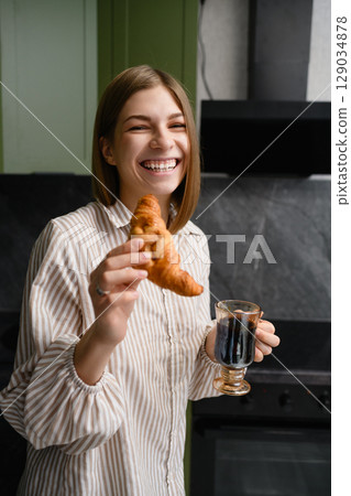 Happy young woman holding croissant and coffee in kitchen. 129034878