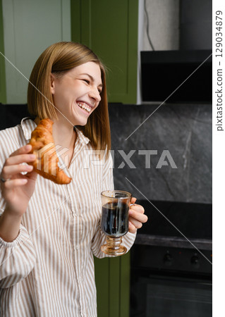 Happy young woman holding croissant and coffee in kitchen. 129034879