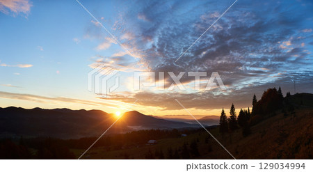 Sun rises behind hills, casting golden glow across sky filled with dramatic clouds. Valley below softly illuminated with mist, enhancing tranquil and picturesque landscape in the morning. 129034994