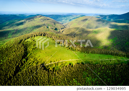 Aerial view of lush, green mountainous landscape. Scene filled with dense forests and rolling hills, bathed in golden light of morning sun. Winding path cuts through greenery, adding to natural beauty 129034995