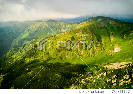 Breathtaking aerial view of green, rugged mountain peaks under dramatic sky. Scene captures contrast between vibrant landscape and moody clouds, with sunlight breaking through. Carpathians, Chornogora Breathtaking aerial view of green, rugged mountain peaks under dramatic sky. Scene captures contrast between vibrant landscape and moody clouds, with sunlight breaking through. Carpathians, Chornogora 129034997
