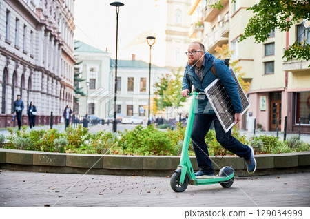 Man rides electric scooter while carrying solar panel. Integration of solar power as sustainable energy source for charging electric scooters, promoting eco-friendly urban transportation. 129034999