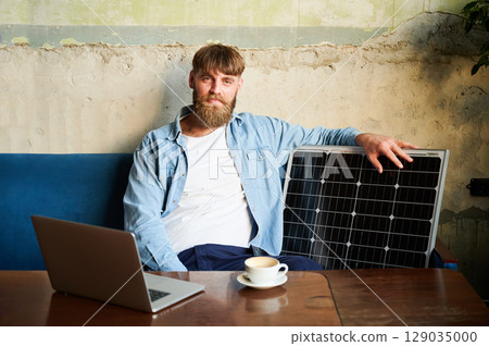 Bearded man works on laptop at wooden table, with cup of coffee. Solar panel nearby, emphasizing sustainable eco-friendly workspace that combines technology and sustainability in cozy environment. 129035000