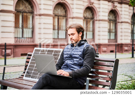 Man sits on wooden bench, working on laptop and smartphone, both connected to solar panel. Urban setting and casual attire highlight modern, sustainable, mobile workspace. Man sits on wooden bench, working on laptop and smartphone, both connected to solar panel. Urban setting and casual attire highlight modern, sustainable, mobile workspace. 129035001
