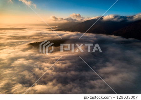 Aerial view of vibrant sunset over white dense clouds bathes in warm golden light, creating dreamlike landscape. Soft shadows play across undulating clouds, while distant landforms peek through mist. Aerial view of vibrant sunset over white dense clouds bathes in warm golden light, creating dreamlike landscape. Soft shadows play across undulating clouds, while distant landforms peek through mist. 129035007