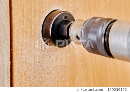 Core cutter with arbor and pilot aligned on battery drill engaged in hole cutting task on door surface viewed close from side angle. Core cutter with arbor and pilot aligned on battery drill engaged in hole cutting task on door surface viewed close from side angle. 129036152
