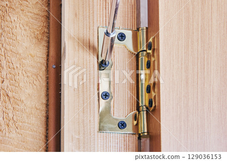Carpenter installing complete door hinge onto wooden door edge, securing metal hinge with screws, detailed view of installation process shown. Carpenter installing complete door hinge onto wooden door edge, securing metal hinge with screws, detailed view of installation process shown. 129036153