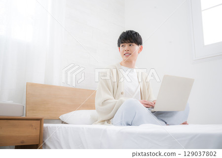 A smiling young man in his 20s studying or working on a computer sitting on his bed. Student or freelancer 129037802