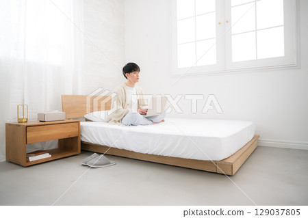 A smiling young man in his 20s studying or working on a computer sitting on his bed. Student or freelancer 129037805