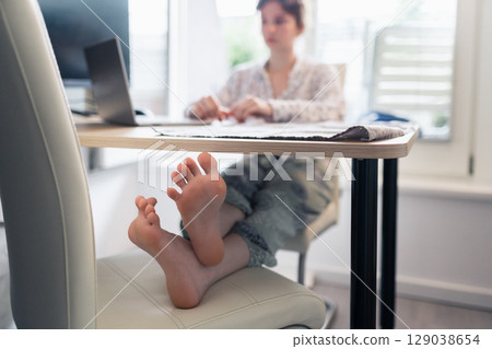 young barefoot woman sitting at table with feet on chair, using laptop 129038654