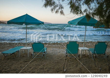 Turquoise beach loungers and umbrellas on a beach with pine trees and stormy sea in the background Turquoise beach loungers and umbrellas on a beach with pine trees and stormy sea in the background 129040249