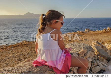 Little 6-year-old girl in striped dress and sunglasses sitting on rocks by the sea 129040414