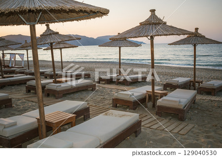 Wooden sunbeds with mattresses and bamboo sun umbrellas on a Greek beach at sunset 129040530