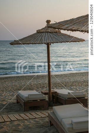 Wooden sunbeds with mattresses and bamboo sun umbrellas on a Greek beach at sunset Wooden sunbeds with mattresses and bamboo sun umbrellas on a Greek beach at sunset 129040604
