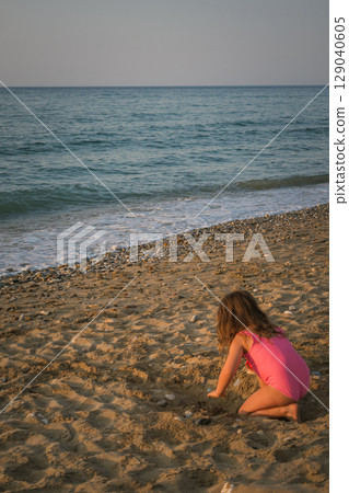 Little girl in a swimsuit playing alone on an empty beach. The ability to entertain oneself without others Little girl in a swimsuit playing alone on an empty beach. The ability to entertain oneself without others 129040605