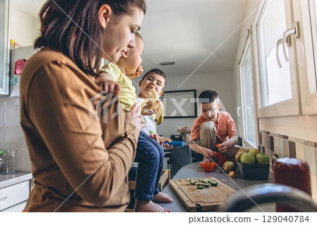 Mother spending time in the kitchen with her children. One child sits on the counter, others play with vegetables. A cozy and lively family moment at home Mother spending time in the kitchen with her children. One child sits on the counter, others play with vegetables. A cozy and lively family moment at home 129040784