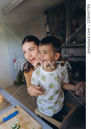 Happy mother hugging her son in the kitchen as they laugh together while preparing fresh cucumber slices. Joyful family bonding during a cooking moment at home. 129040789