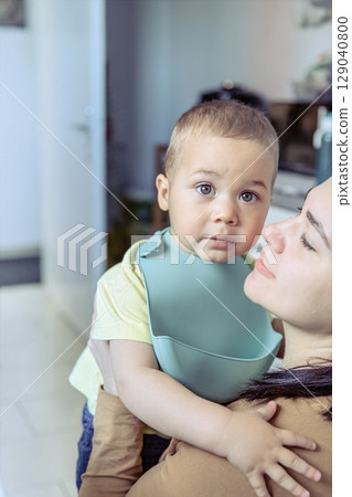 Mother gently holds her toddler wearing a silicone bib in a kitchen setting. The child looks at the camera with a calm and curious expression 129040800