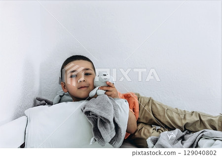 Little boy lying in bed with a soft toy, relaxing under a blanket in a bright white room Little boy lying in bed with a soft toy, relaxing under a blanket in a bright white room 129040802
