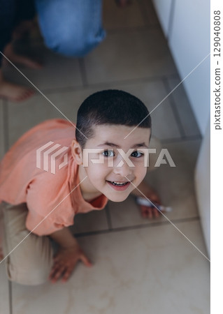 Happy young boy crouching on the kitchen floor, smiling up at the camera while holding a small object 129040808